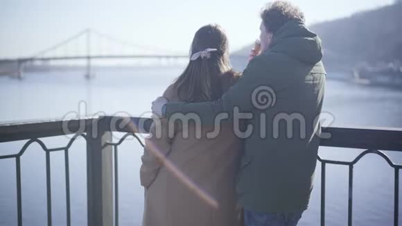 在河畔拥抱女友和吃冰淇淋的快乐年轻白种人男人的后景恋爱中的情侣视频的预览图