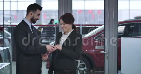 兴奋的高加索女人从男性经销商那里得到新的红色汽车钥匙并做出胜利手势的肖像满意视频的预览图
