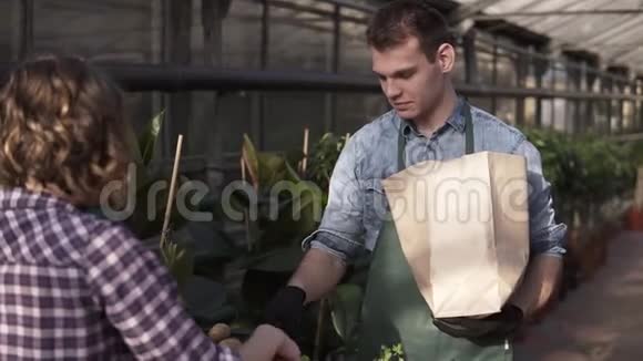 白种人穿绿色围裙的高大推销员正在温室市场上给顾客放新鲜沙拉胡萝卜装在纸袋里视频的预览图