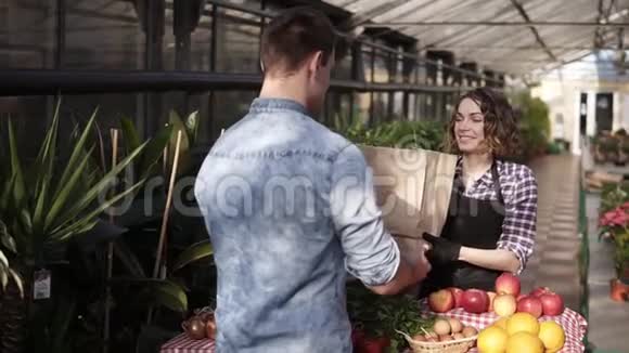 穿着黑色围裙的开朗的女售货员在温室市场上给顾客提供纸袋中的新鲜蔬菜微笑的女人视频的预览图