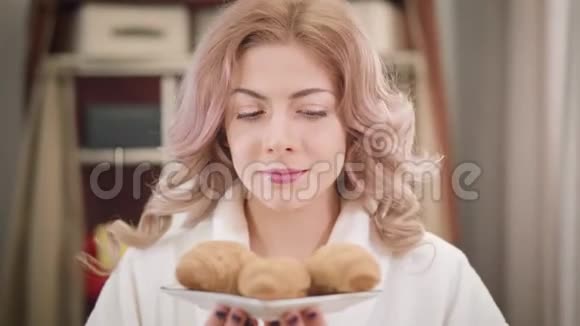 美丽的白种人女孩闻着美味的牛角面包的肖像并向相机展示盘子快乐的年轻女子视频的预览图