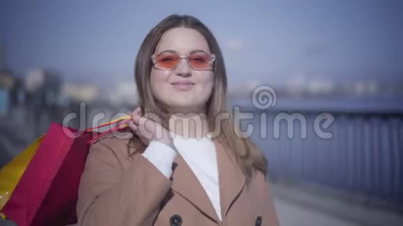 一位戴着太阳镜带着购物袋在户外摆姿势的笑容满面的白人胖乎乎女孩的特写肖像快乐的年轻女子视频的预览图