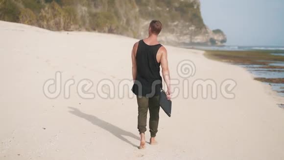 后景手拿瑜伽垫的孤独男子沿着沙滩漫步视频的预览图