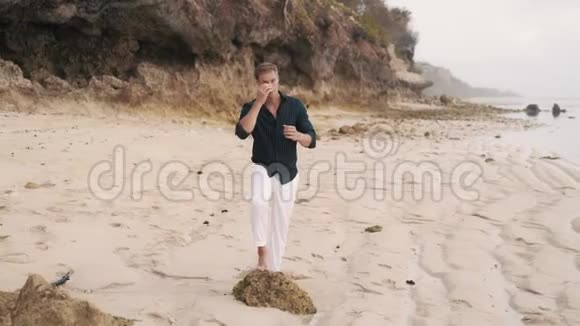 穿着白色裤子和黑色衬衫的帅哥站在海滩上看着大海视频的预览图