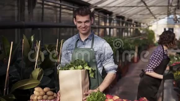 年轻的白种人穿着围裙手里拿着一个棕色的纸袋里面装着新鲜的蔬菜和蔬菜视频的预览图