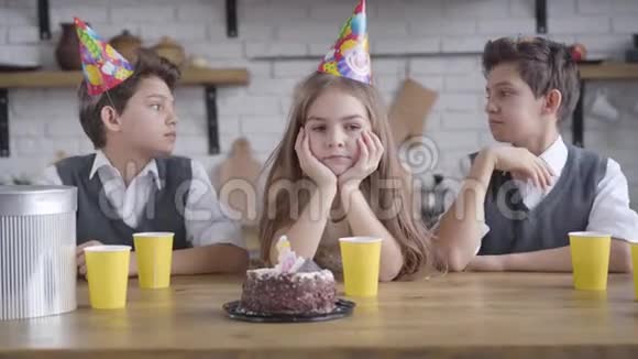 一幅美丽的小悲伤女孩坐在桌子旁双胞胎兄弟戴着派对帽的肖像高加索少年视频的预览图