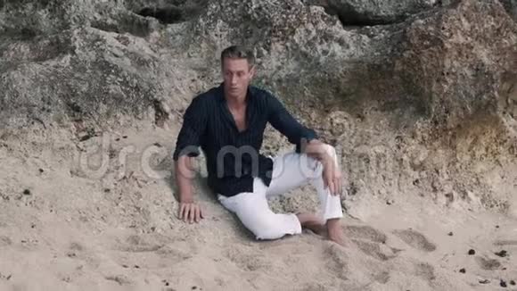 穿着白色裤子和黑色衬衫的野蛮人坐在岩滩上看着摄像机视频的预览图