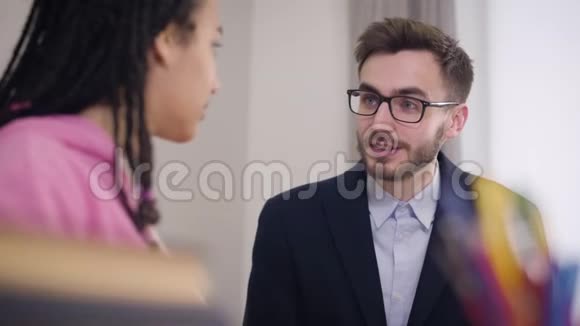 年轻的黑发白种人男人灰色的眼睛和非裔美国女孩说话穿西装戴眼镜的优雅老师视频的预览图