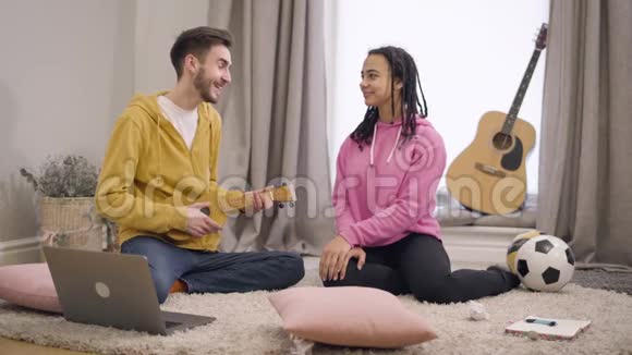 年轻微笑的白种人男孩唱小夜曲为他的非裔美国女友演奏四弦琴恋爱中的情侣视频的预览图
