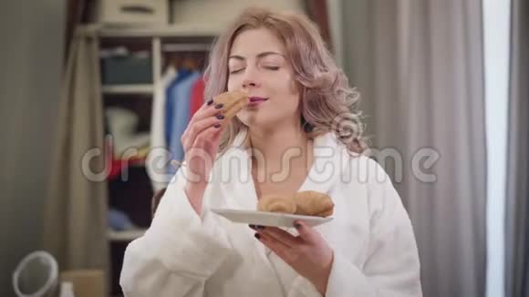 美丽的白种人女人穿着白色浴袍闻着美味的牛角面包咬着它的肖像可爱的女士视频的预览图