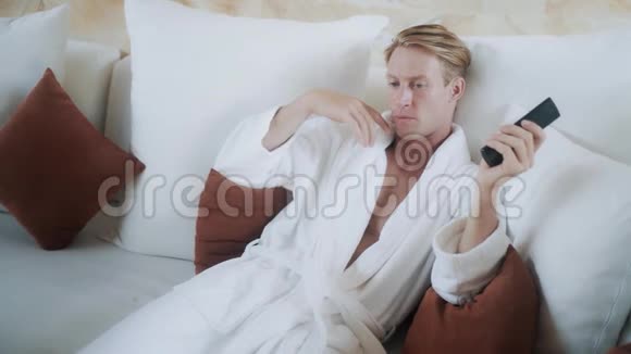 穿着浴袍的金发男人在舒适的床上打开电视视频的预览图