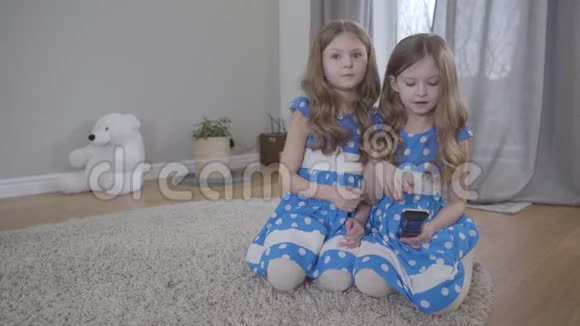 迷人的黑发白种人双胞胎姐妹的肖像切换频道与遥控器穿蓝色衣服的快乐孩子视频的预览图