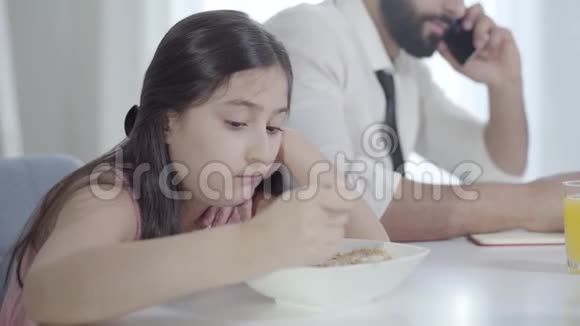 令人沮丧的中东漂亮女孩的肖像回首看着无法辨认的父亲在早餐时打电话视频的预览图
