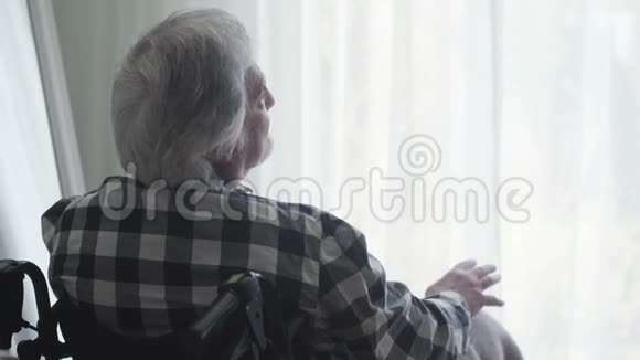 坐在轮椅上的孤独老人坐在大窗户前的近景窗帘和视频的预览图
