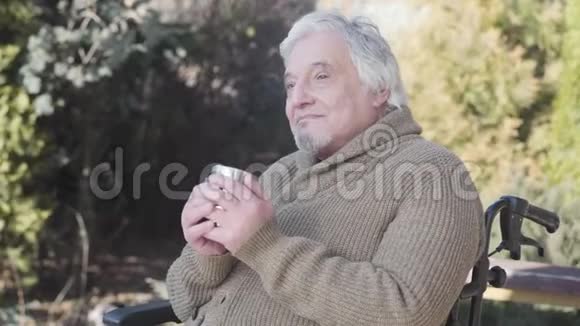 侧面的肖像老人白种人喝热茶坐在轮椅户外老年男性退休人员视频的预览图