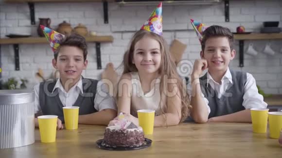 慢动作两个白种人双胞胎男孩和戴着派对帽的可爱女孩端着生日蛋糕坐在桌子旁看着视频的预览图