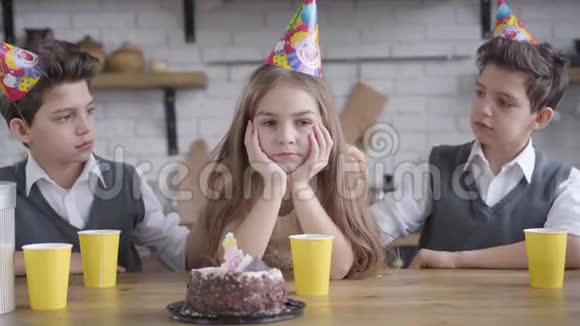 双胞胎兄弟颤抖着肩膀抚摸着戴着派对帽的悲伤白种人女孩的头发坐在桌子旁的儿童心烦意乱视频的预览图