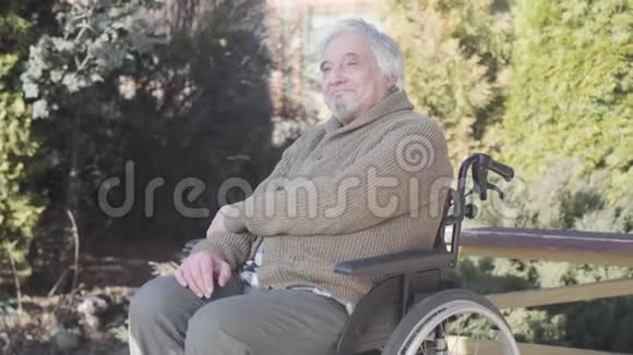 微笑的老白种人残疾人坐在轮椅上向远处看的肖像穿暖和衣服的老年退休人员视频的预览图