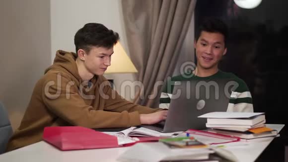两个智能微笑的大学生坐在桌子旁用笔记本电脑快乐的白人和亚裔青少年视频的预览图