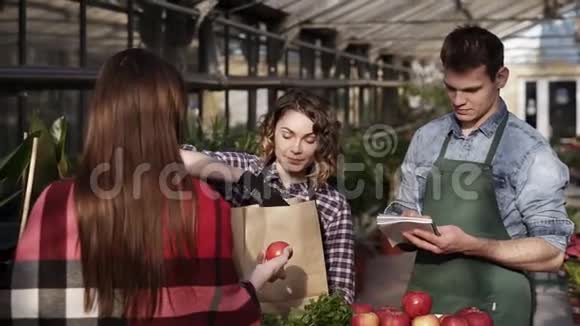穿围裙的欧洲女售货员正在温室里给顾客提供有机食品女人正在包装蔬菜水果视频的预览图