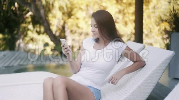 穿着白色t恤的女孩在泳池沙发上打电话上网视频的预览图