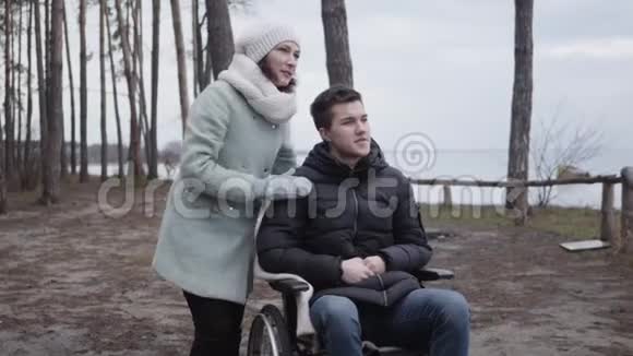 积极的白人男子坐轮椅成年妇女在河岸的森林里眺望无效投标或视频的预览图