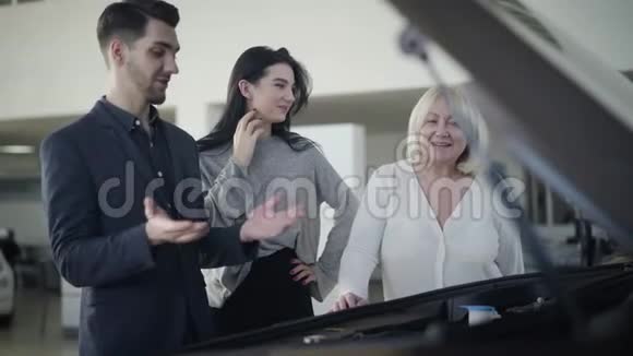 年轻的白种人白种人和资深的金发母亲和英俊的经销商交谈站在旁边打开汽车罩视频的预览图