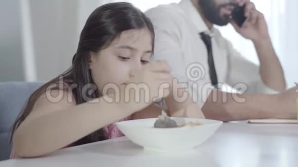 一个可爱的中东小女孩用勺子混合谷物就像一个无法辨认的忙碌男人在电话中交谈视频的预览图