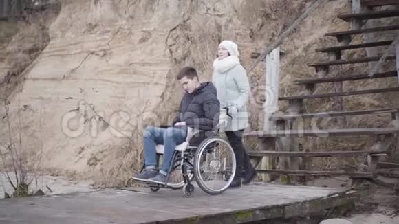 年轻的白人妇女在码头上滚动轮椅带着悲伤的残疾男孩有思想的人度过秋日的画像视频的预览图