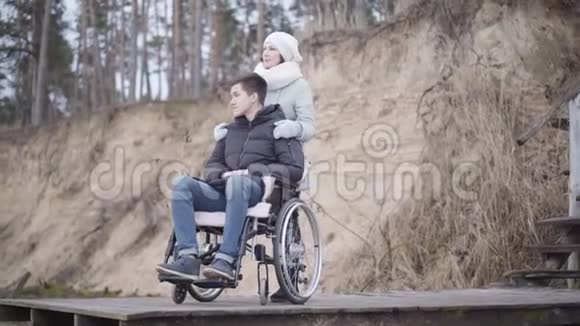 与母亲坐在轮椅上的高加索男孩或无效的温柔可怜的残废人在户外度秋日视频的预览图