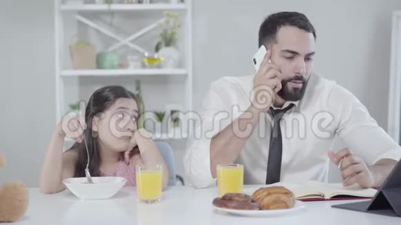 中东小女孩看着父亲在早上打电话忙碌的男人和无聊的早餐视频的预览图