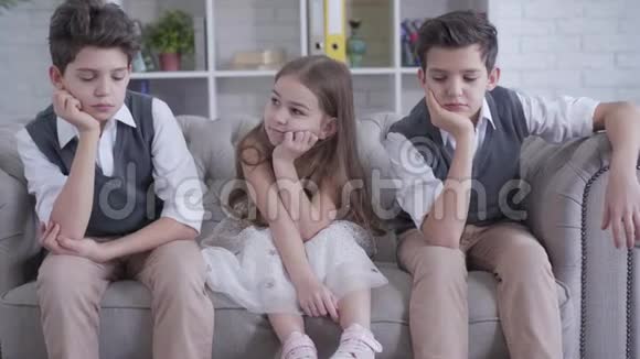 白种人双胞胎兄弟坐在沙发上两边都是欢快可爱的女孩无聊儿童在室内休息的肖像视频的预览图