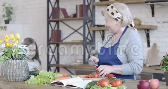 积极的金发退休者切割番茄因为小黑发女孩来帮助她做饭快乐的肖像视频的预览图