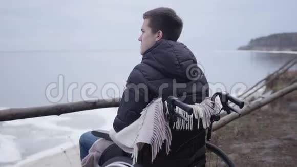 沮丧的白人男孩坐在轮椅上独自在河岸度过秋日可怜的残疾青年画像视频的预览图