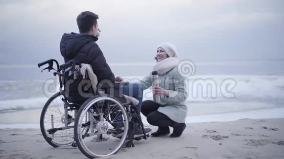 坐在轮椅上的年轻白种人男孩在海边喝茶和坐在舱里的美女交谈视频的预览图
