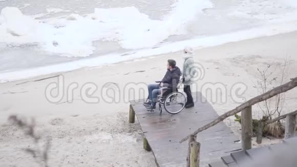 坐在轮椅上的白种人残疾男孩和站在码头上看着结冰的河流的无效投标青年残疾人视频的预览图