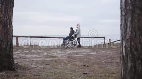 在户外轮椅上站在年轻男子旁边的白人女人的极长镜头残疾男孩和残疾儿童的侧视图视频的预览图