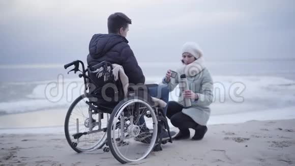漂亮的白种人女人从热水瓶里倒茶然后把它送给轮椅上的残疾男孩微笑无效视频的预览图