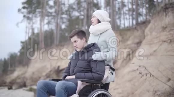 年轻英俊的残疾男孩坐在轮椅上向下和向上看户外一位严肃的高加索女人抱着他的肩膀视频的预览图