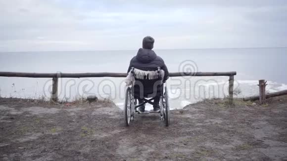 黑黑肤色白种人坐在河岸上的轮椅后景悲伤的残疾男孩望着远方想着视频的预览图
