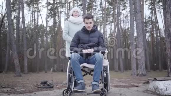 成年白种人妇女扶着轮椅上悲伤残疾男孩的肩膀说话母亲或无效投标视频的预览图
