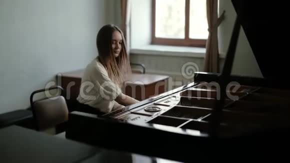 一位年轻的钢琴家长发在窗户附近的大钢琴上弹奏音乐视频的预览图