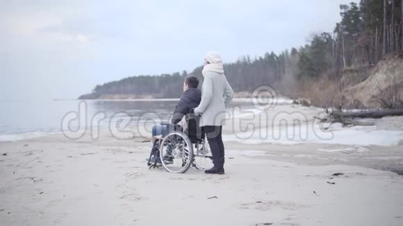 在沙质河岸上一个有思想的白人妇女和残疾的儿子站在轮椅上母亲肖像视频的预览图