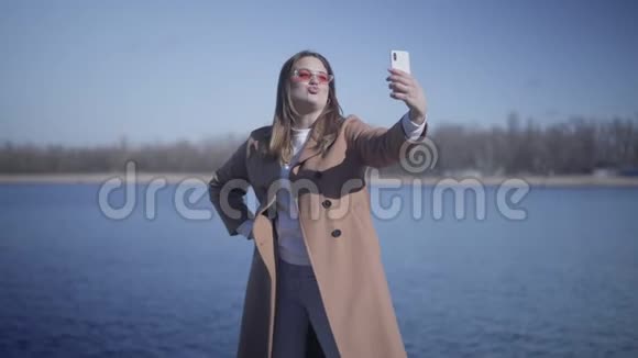 正面年轻女性在户外自拍相机上发送空气亲吻和鬼脸的肖像快乐的白人女孩视频的预览图