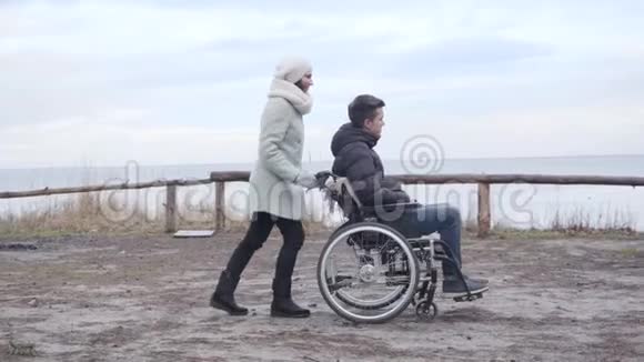 河岸边有残疾男孩的成年白种人妇女滚动轮椅的侧景微笑的镜头视频的预览图