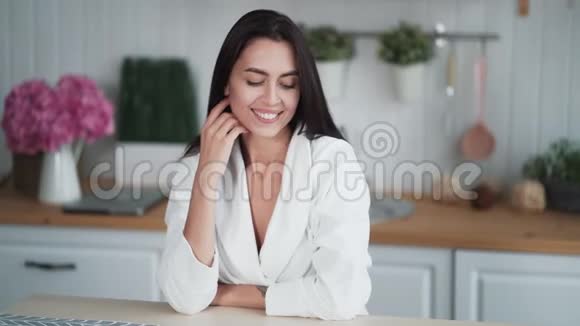一幅可爱的年轻女子坐在家里的厨房里看着镜头微笑视频的预览图