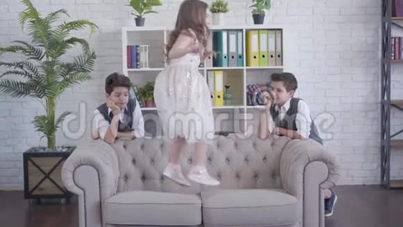 精疲力竭的孪生兄弟站在沙发的两边看着他们欢快的妹妹在沙发上跳跃累了视频的预览图