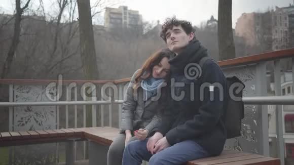 快乐的白人夫妇坐在公园的长凳上微笑女孩抱着头男孩的肩膀和男人看着视频的预览图