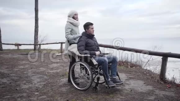 年轻积极的白人妇女触摸轮椅上残疾男子的肩膀并指向别处的肖像强国视频的预览图
