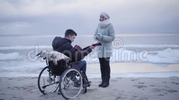 年轻的白种人残疾男子在河岸弹吉他为母亲或无效的投标坐轮椅的男孩视频的预览图
